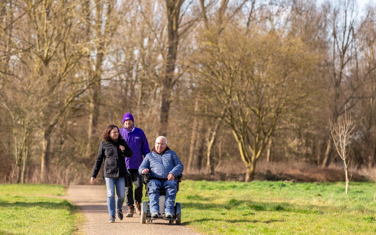 Wheelchair user with two people walking along a footpath surrounded by winter trees.