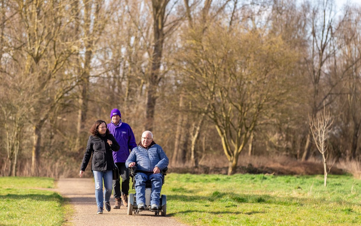 Wheelchair user with two people walking along a footpath surrounded by winter trees.