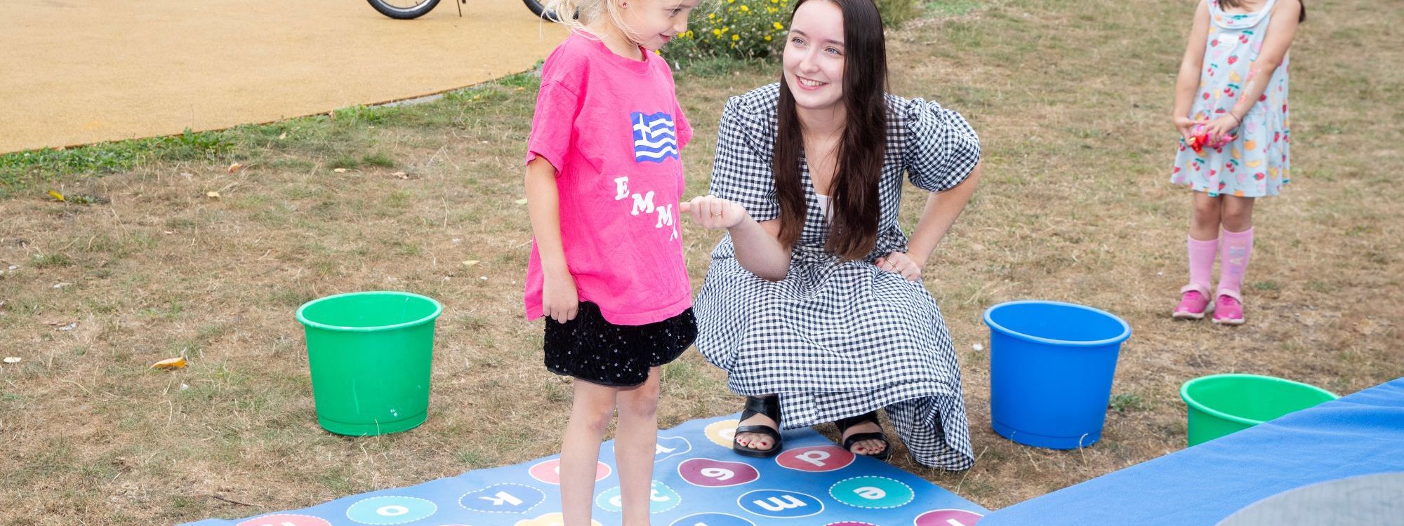 Young child standing on a mat with many numbers on, playing a maths game with a tutor in a park.