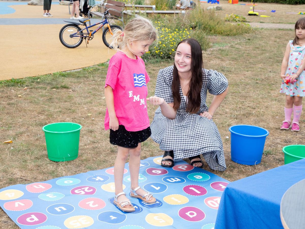 Young child standing on a mat with many numbers on, playing a maths game with a tutor in a park.